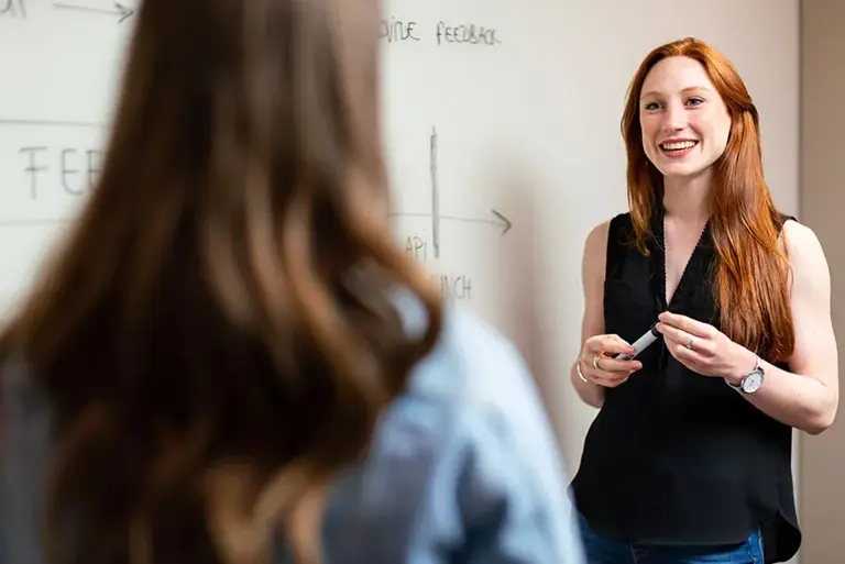 Two females workers discuss the issue they have discovered on the large whiteboard behind them