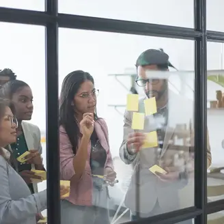 Four office workers are seen through a window into a conference room where they are working on a series of post-it notes on the glass.