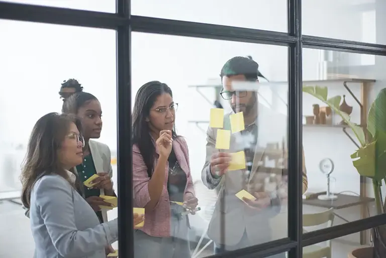 Four office workers are seen through a window into a conference room where they are working on a series of post-it notes on the glass.