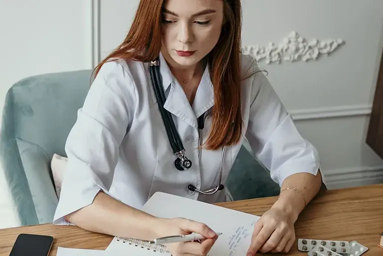 Doctor sits a desk and writes notes in her paper notebook