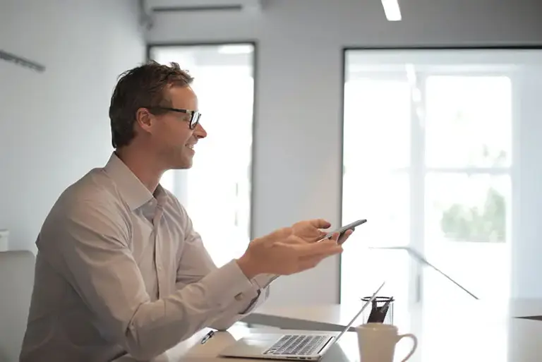 Office worker, sits smiling at a office desk, in front ofa n open laptop in bright glass walled office