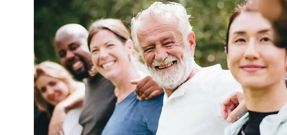 A line of people of all ages and diverse ethnic backgrounds smile to the camera while they all have their arms around each other's backs