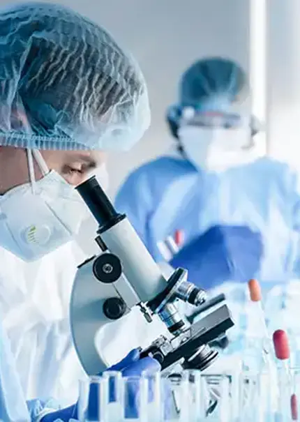 Two masked scientists in a lab, the one in the background takes note with the closer one looks into a microscope