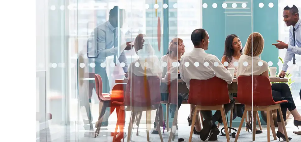 At a brightly lit all glass meeting room, on man stands and addresses the four seated delegates 