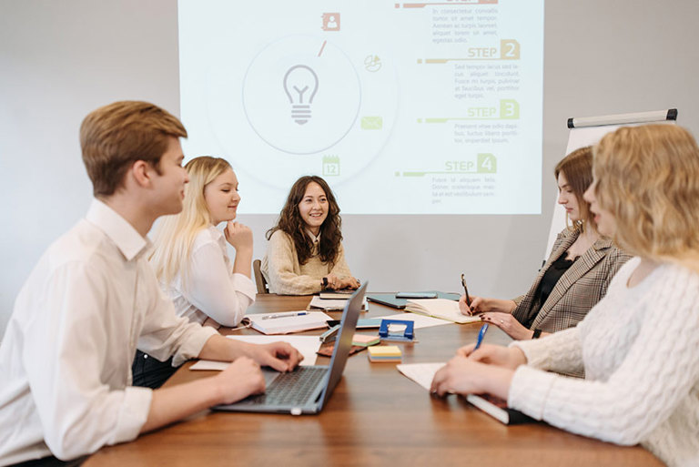 Team of office workers sit at a large table with the presentation projected on the rear wall