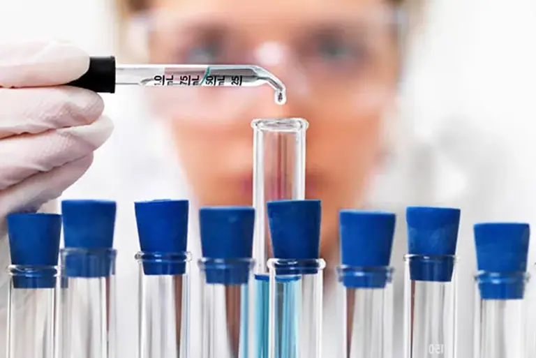 Close up of a rack of test tubes with a scientist adding a single drop from a pipette into the middle test tube.