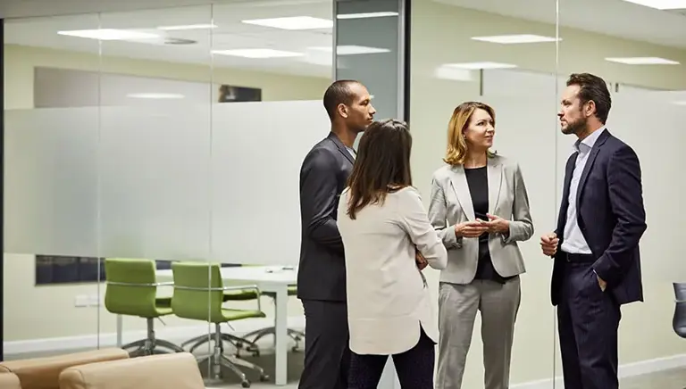 Four office workers stand talking in front of a set of glass meeting rooms