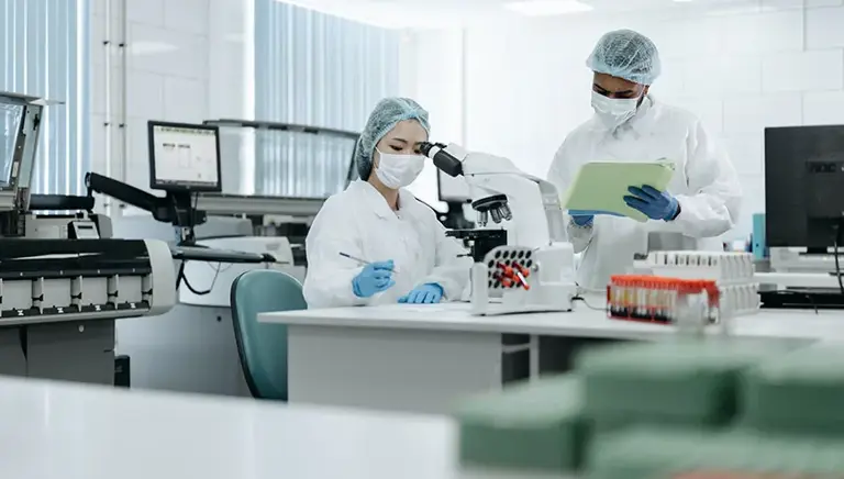 Two scientist are working in lab. One is seated and making notes after looking down her microscope, while her colleague stands nearby and he makes notes on a clipboard.