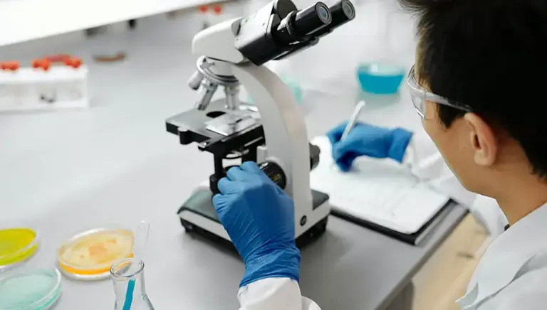 Scientist sits working at a microscope in a lab, writing notes.  