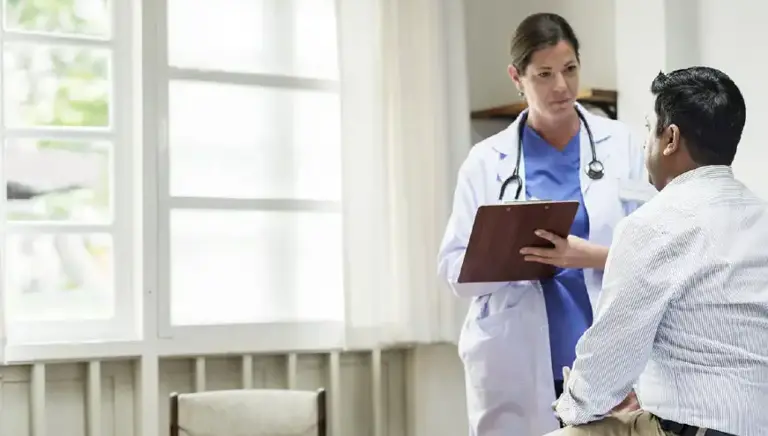 Doctor with a clipboard stands interviewing a patient sitting on a bed in a hospital setting